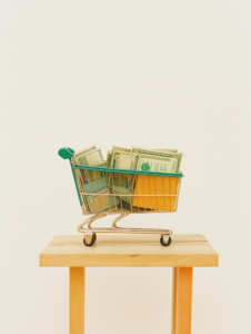 A scale balancing an overflowing shopping cart against a handful of dollar bills