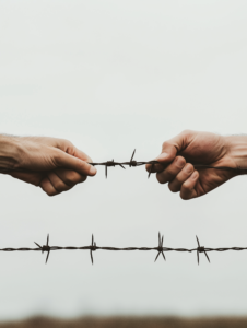 Two hands stretch towards each other from opposite sides of a barbed wire fence, illustrating the desperate desire to connect