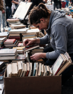 A person digging through a box of old books at a flea market, searching for hidden literary treasures